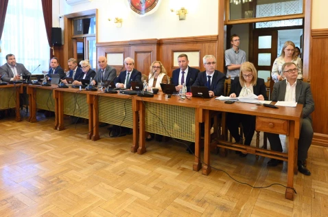 Radni Powiatu i skarbnik siedzą przy biurkach w sali sesyjnej. W tle stoją pracownicy Starostwa.