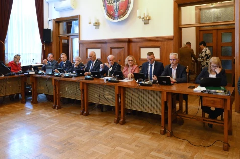 Radni Powiatu i skarbnik siedzą przy biurkach w sali sesyjnej. W tle stoją 2 pracownicy Starostwa.