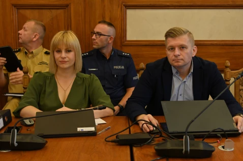 Radna Katarzyna Stadnik i radny Łukasz Krupa siedzą przy laptopach w sali sesyjnej. Za nimi siedzi policjant i strażak.